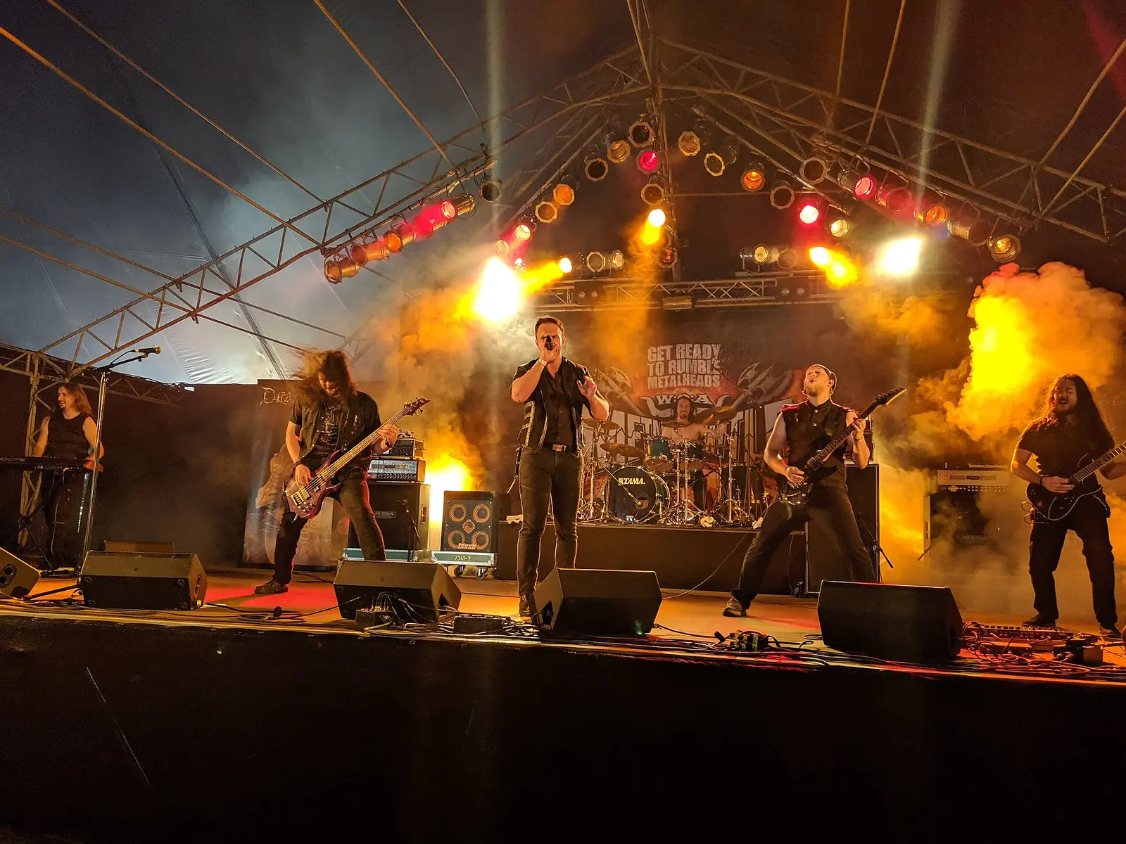 A band performing on a stage with coloured lights and smoke at Wacken Open Air in 2019